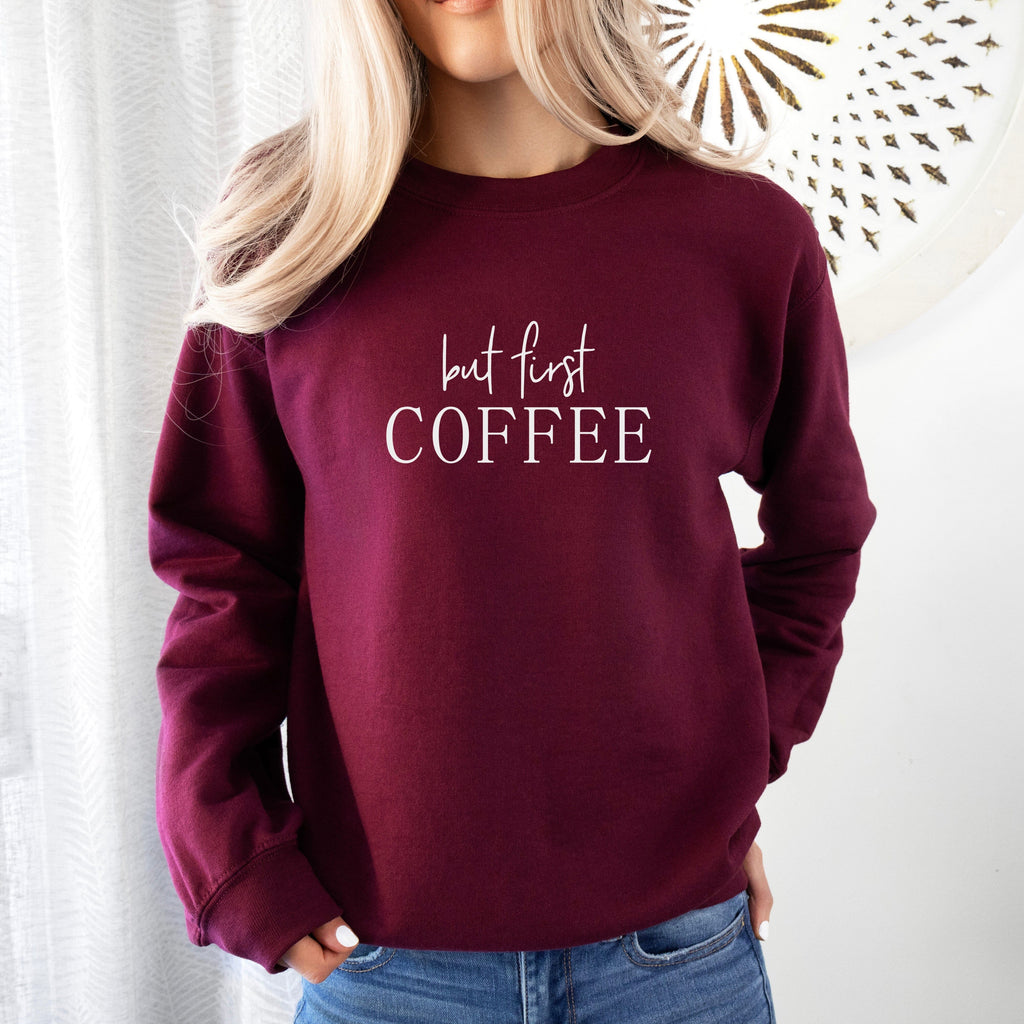 Maroon sweatshirt with 'but first COFFEE' text worn by a person, against a white background.